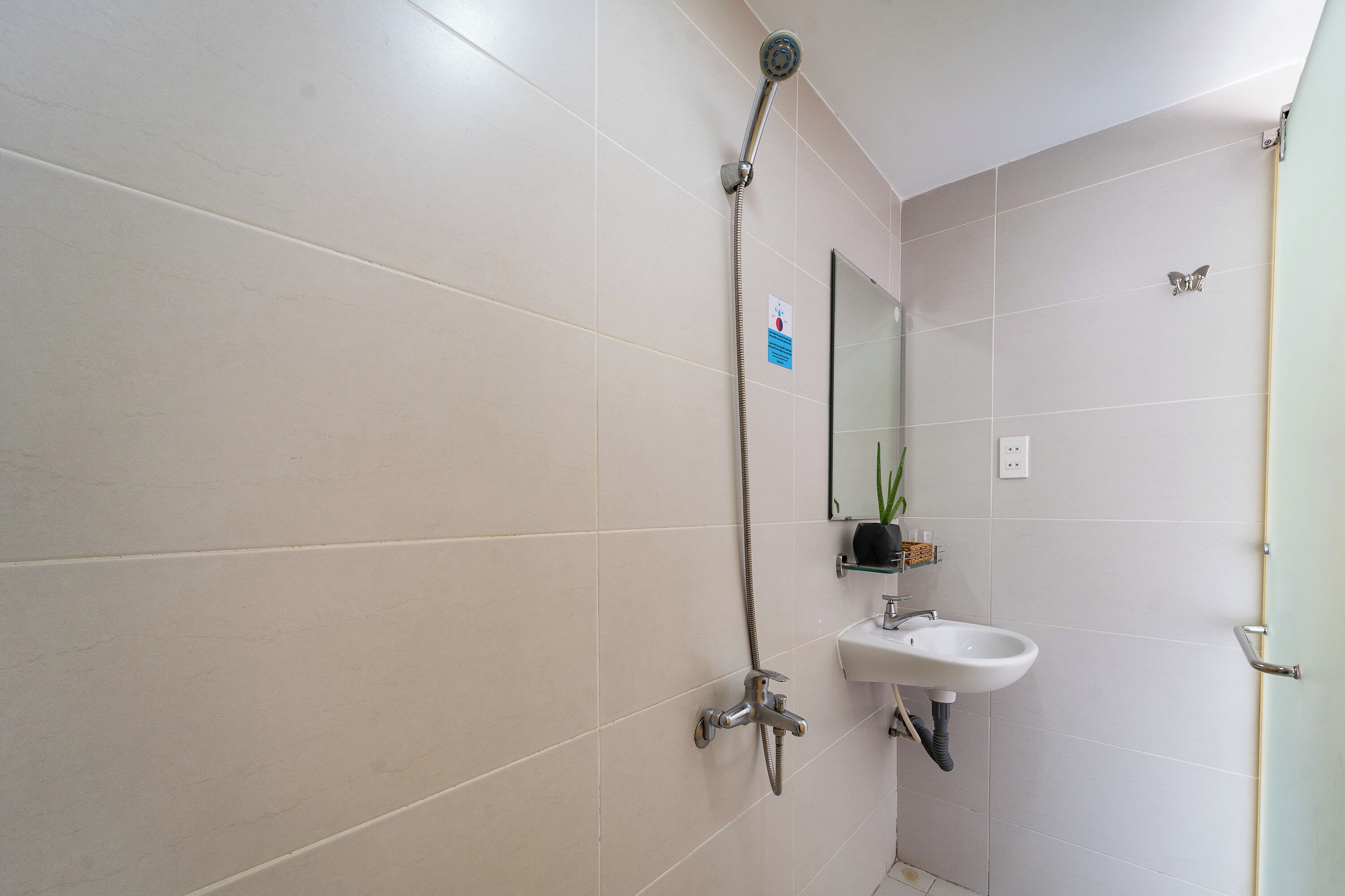 A bathroom with white tiled walls, a shower with a handheld showerhead, a sink with a mirror above it, and a small shelf with toiletries.