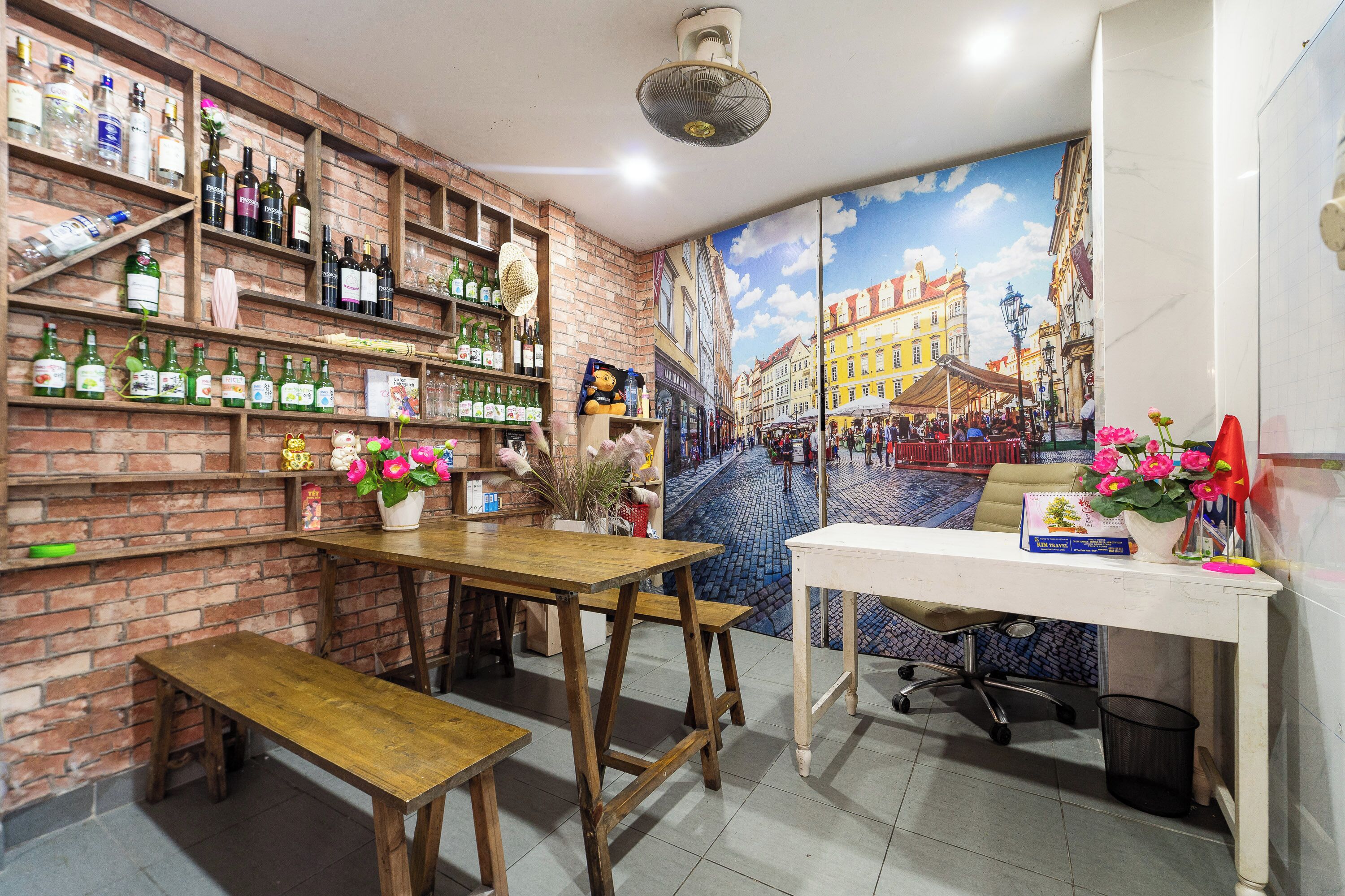A cozy bar area with wooden shelves stocked with various bottles, a wooden table with benches, and a mural of a street scene on the wall.