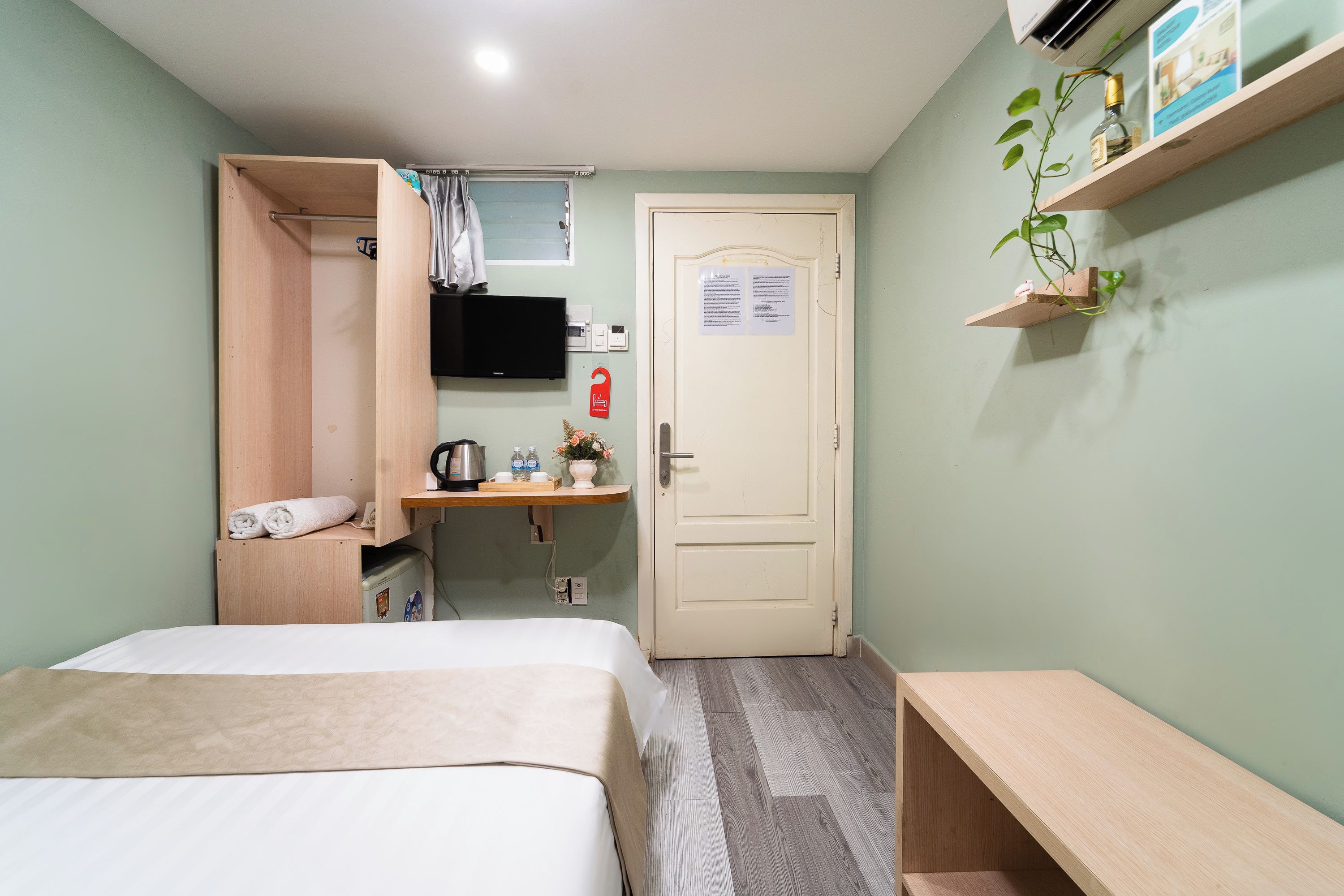 A compact hotel room with a bed, a small desk with a kettle, a television, and a door with a notice.