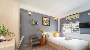 A hotel room with a bed, a desk, a chair, a small table with a vase of flowers, a wall-mounted TV, and an air conditioning unit.