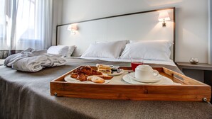 Cama bem arrumada com lençóis brancos, um tabuleiro com pastelaria e uma chávena de café, e um abajur.