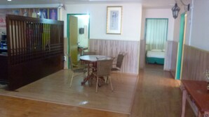 Una habitación con piso de madera, una mesa de comedor con sillas, una cama con colcha verde y un cuadro enmarcado en la pared.