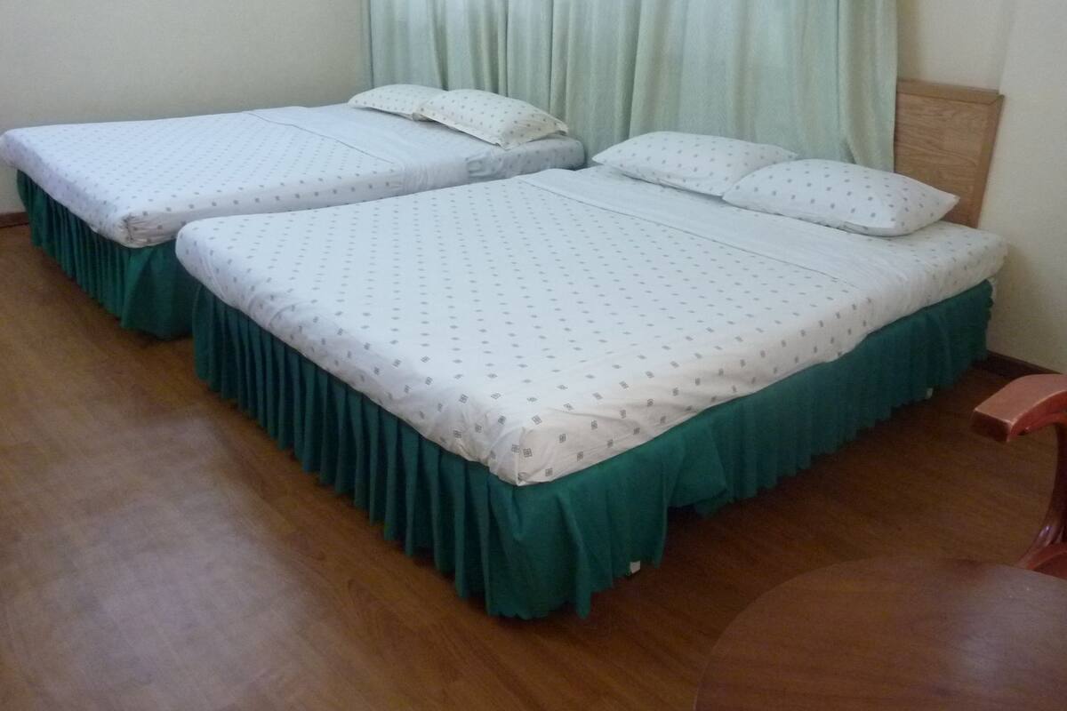 Two single beds with green bed skirts, a wooden chair, and a wooden table in a room with a wooden floor.