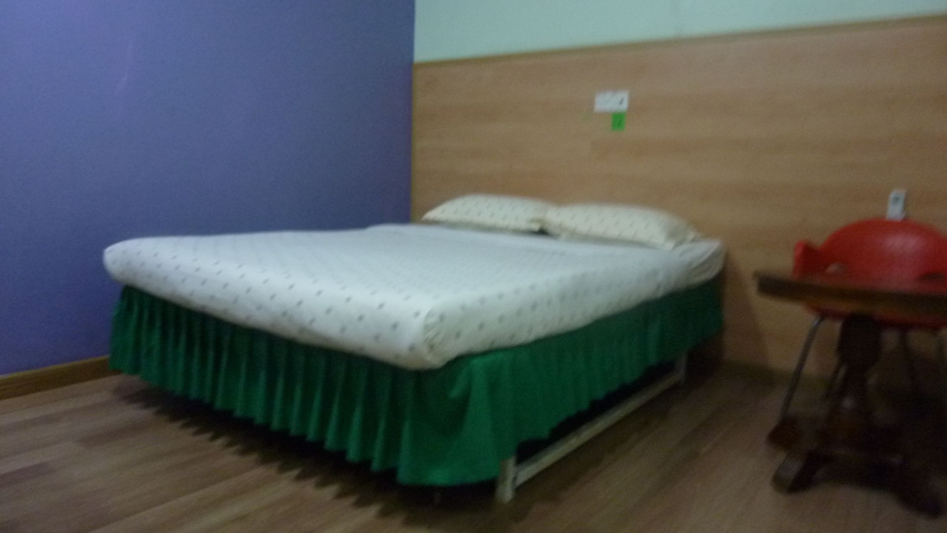 Uma cama de solteiro com saia verde e roupa de cama branca em quarto com piso de madeira e parede roxa.