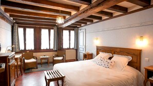 Une chambre à coucher avec un grand lit, une tête de lit en bois, deux tables de chevet, une commode, une chaise, une petite table et une fenêtre avec des rideaux.
