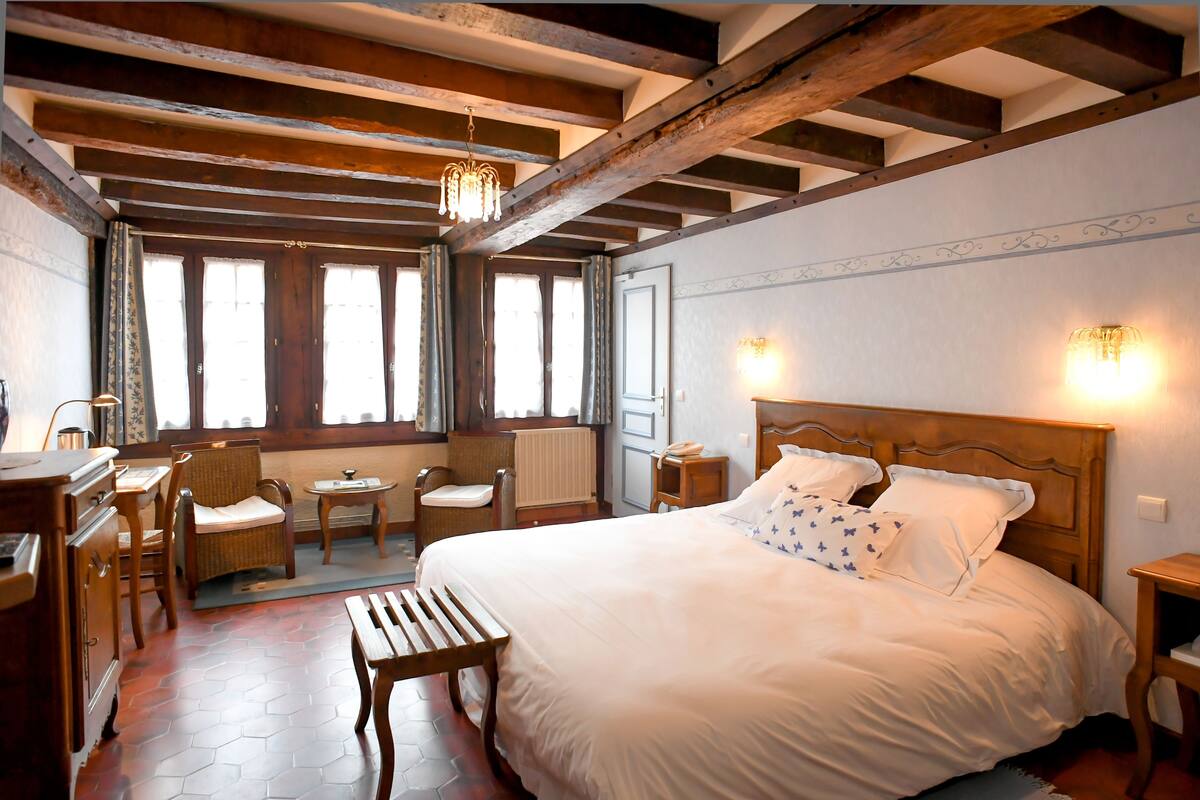 Une chambre à coucher avec un grand lit, une tête de lit en bois, deux tables de chevet, une commode, une chaise, une petite table et une fenêtre avec des rideaux.