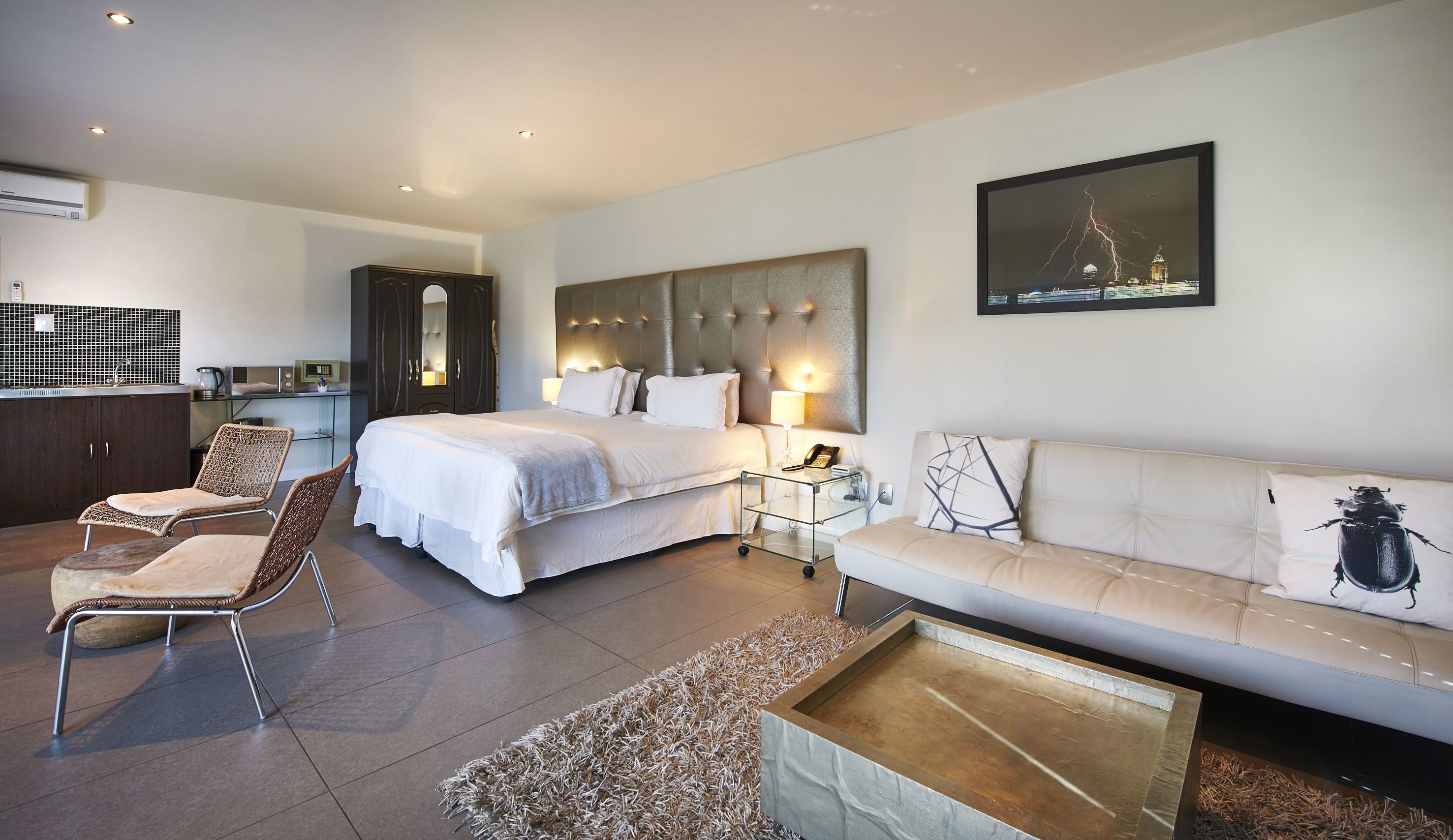 A modern hotel room with a large bed, a sofa, a dining area, and a kitchen.