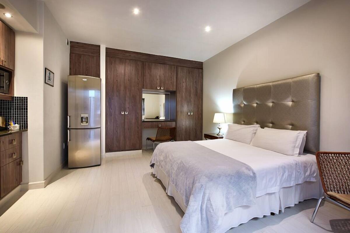 A modern hotel room with a large bed, a built-in wardrobe, a refrigerator, and a seating area with a chair and table.