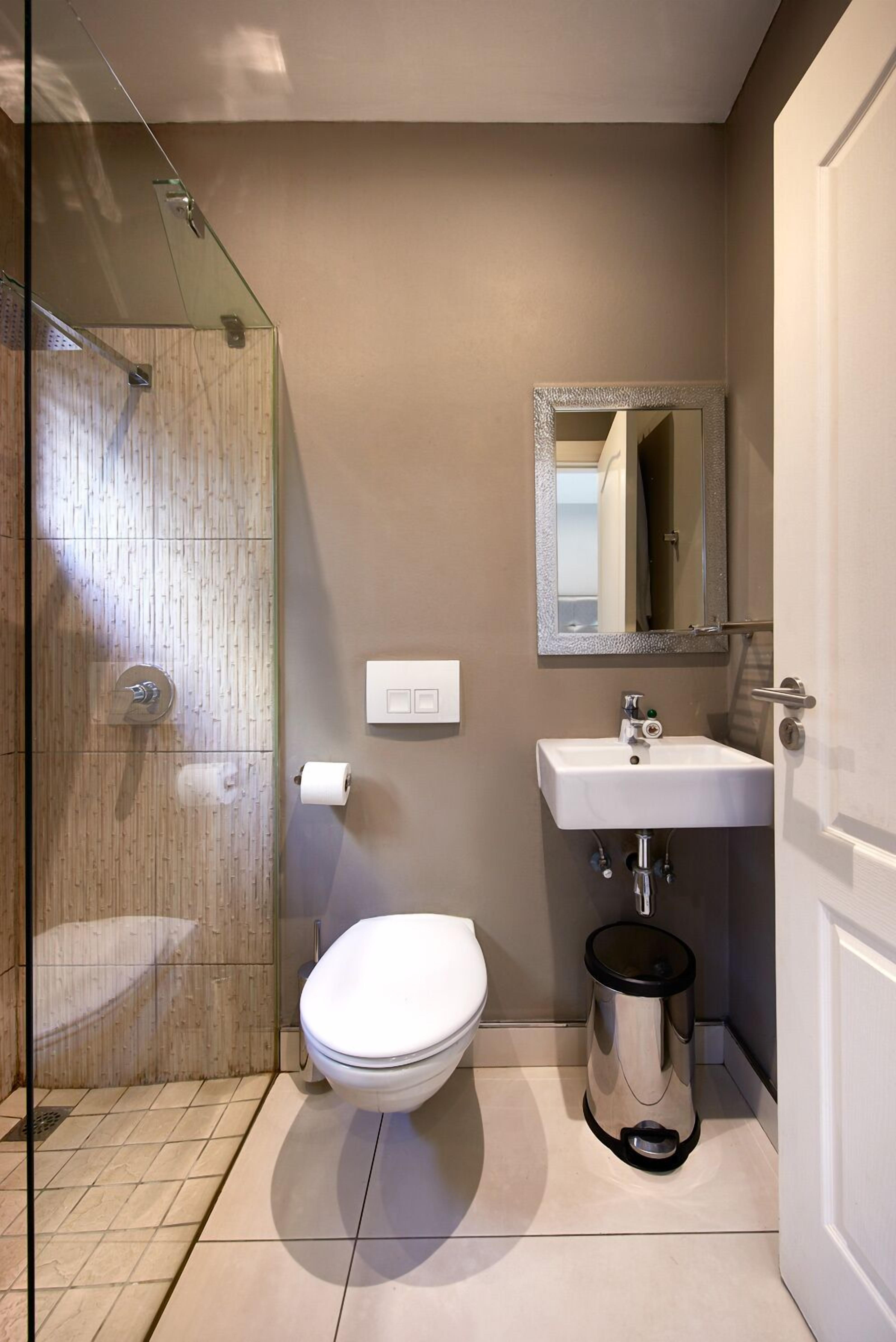 A bathroom with a glass shower enclosure, a white toilet, a sink with a mirror, and a trash bin.