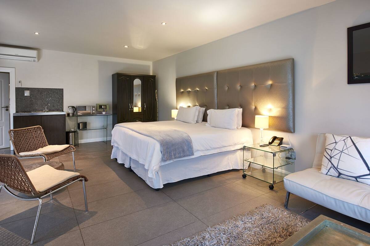 A modern hotel room with a large bed, a sofa, a chair, a small table with a lamp, and a TV on the wall.