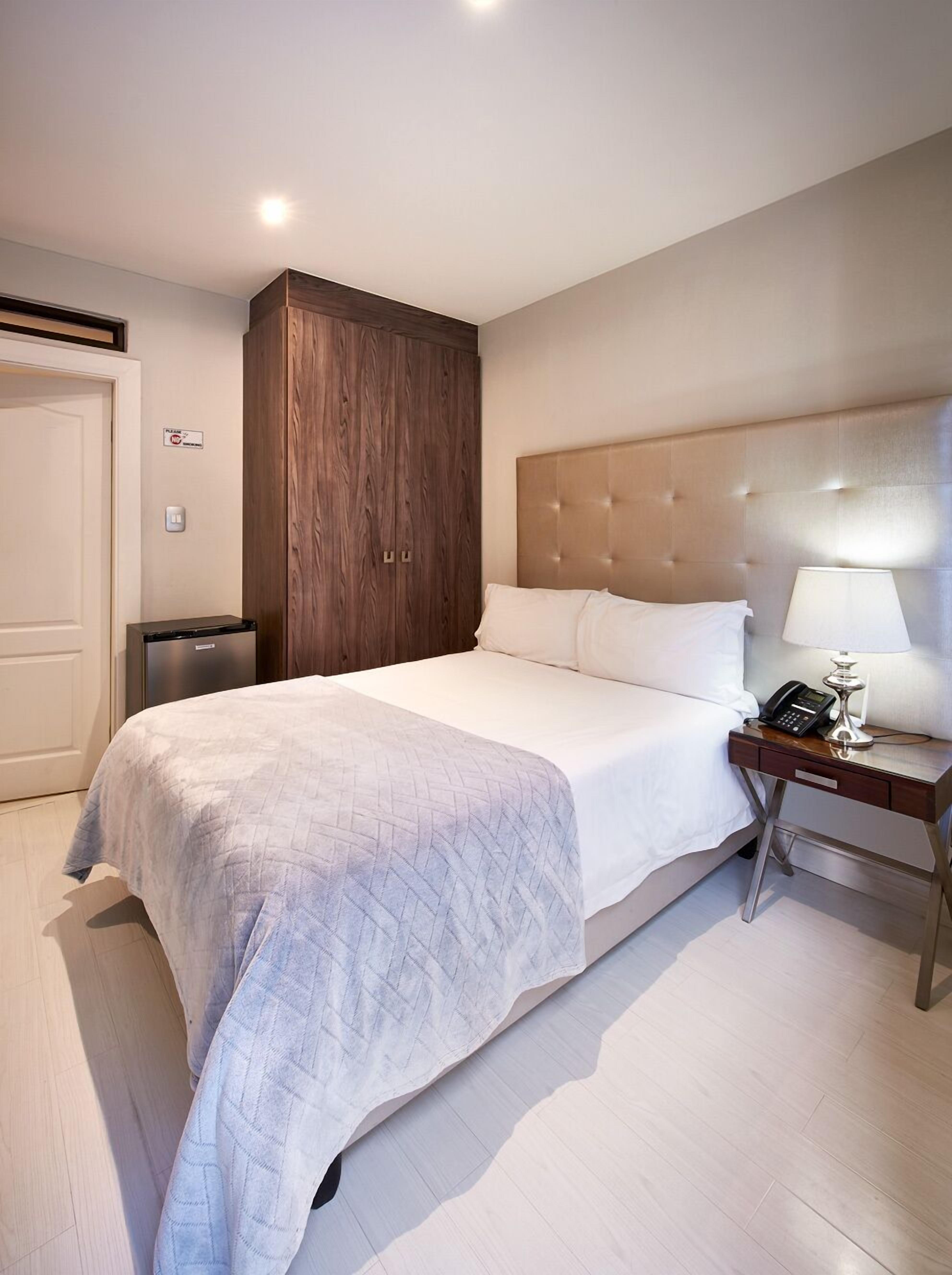 Quarto de hotel com cama, mesa de cabeceira, guarda-roupa de madeira e uma porta.