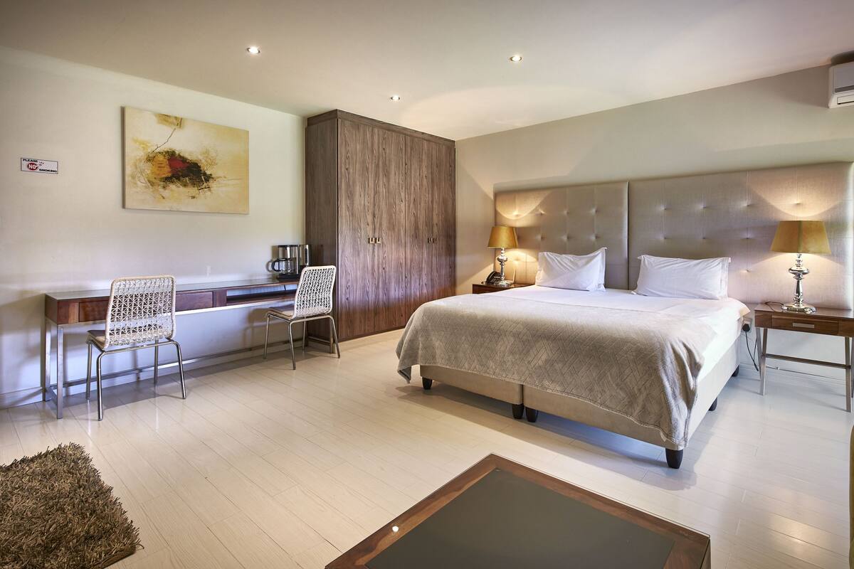 A modern hotel room with a bed, desk, chair, and a large artwork on the wall.