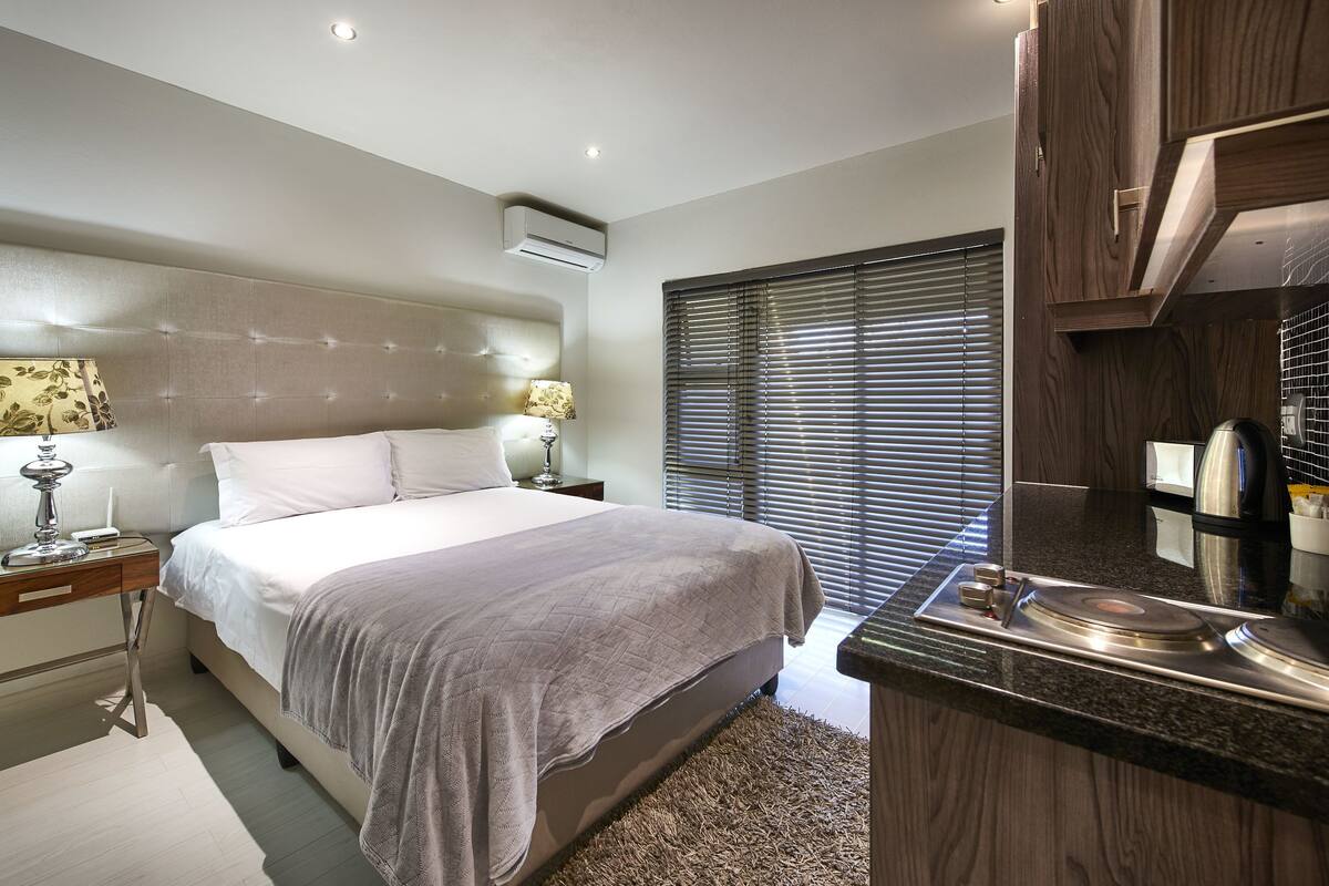 A modern hotel room with a large bed, bedside lamps, a wooden cabinet, and a kitchen area with a coffee maker.
