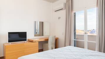 A hotel room with a bed, a TV on a wooden dresser, a desk with a chair, a window with curtains, and an air conditioning unit.