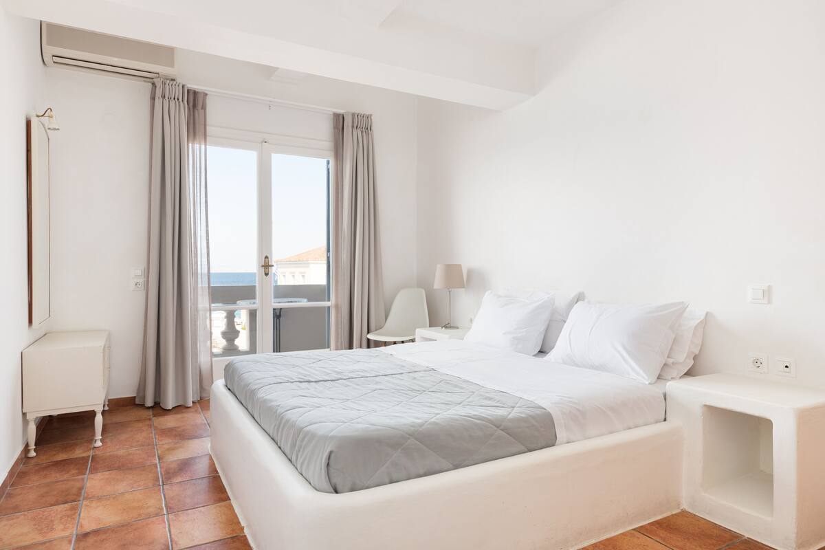 A hotel room with a large bed, a desk, a chair, and a view of the sea through the window.