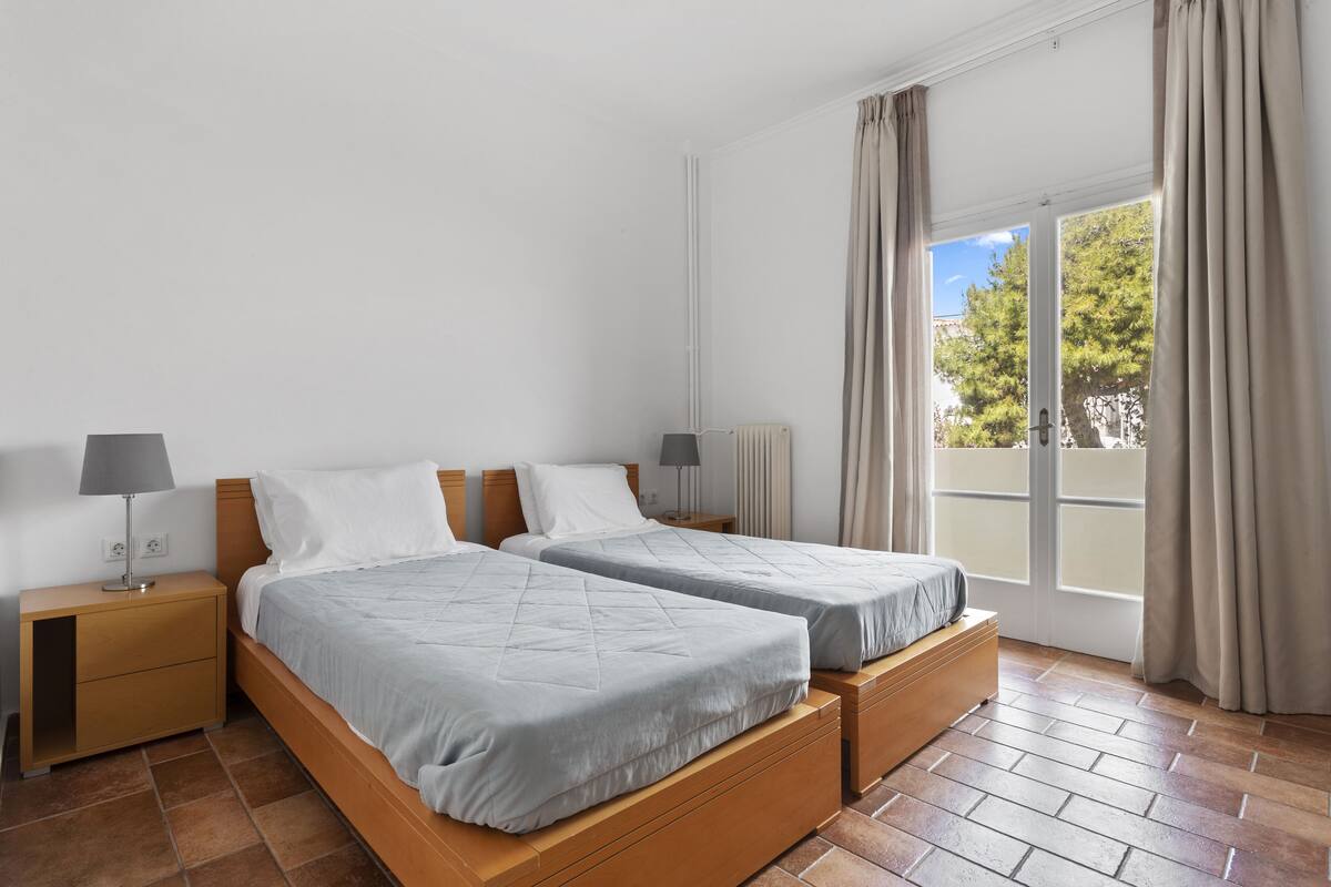 A double room with two beds, a nightstand with a lamp, a window with curtains, and a view of trees.