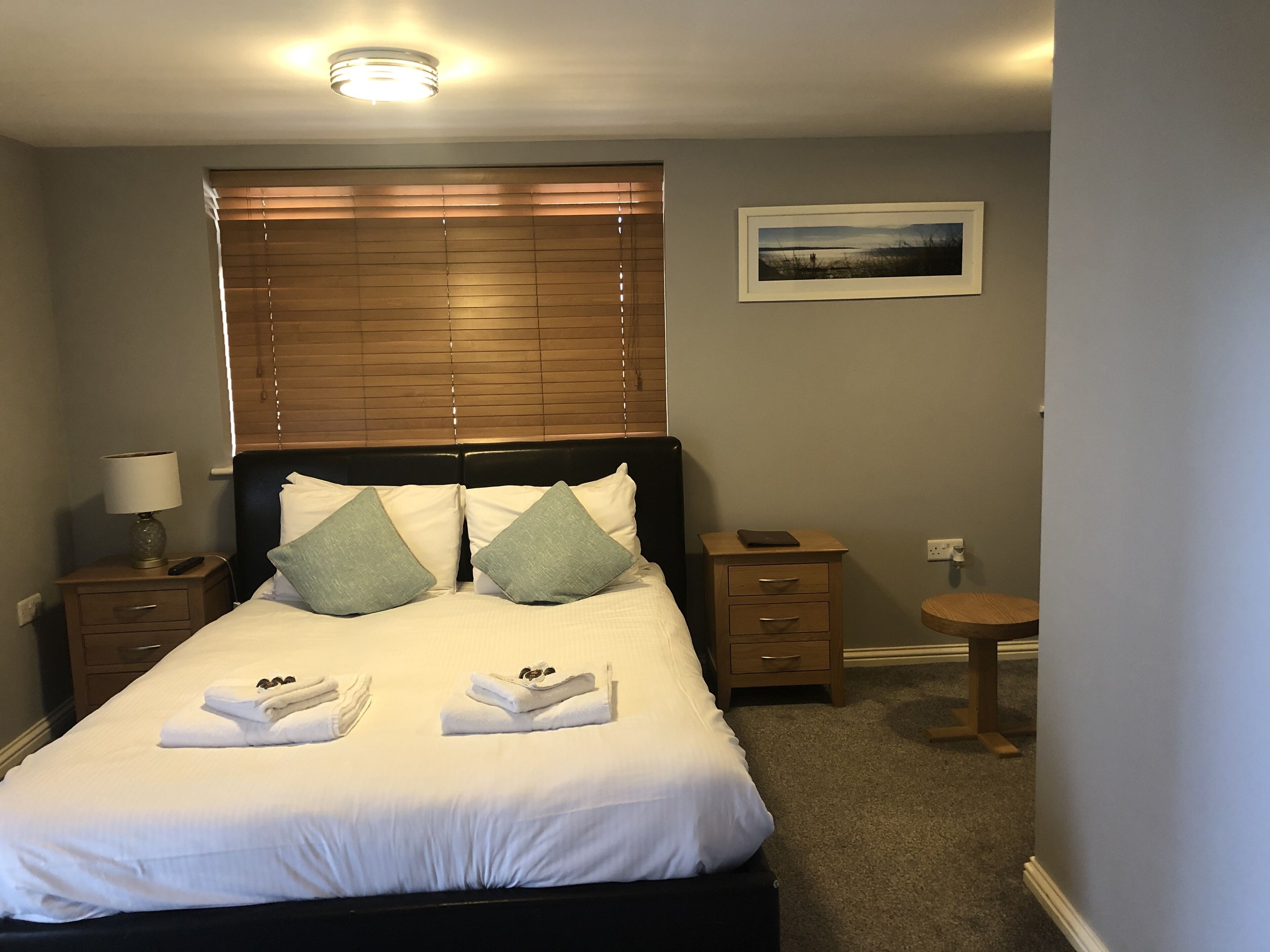 A neatly made bed with white linens and two pillows, a bedside table with a lamp, a small wooden nightstand, and a framed picture on the wall.