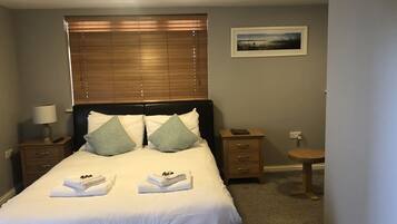 A neatly made bed with white linens and two pillows, a bedside table with a lamp, a small wooden nightstand, and a framed picture on the wall.