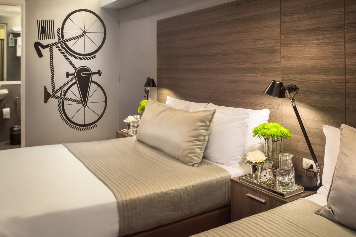 A hotel room with two beds, a wooden headboard, bedside lamps, a clock, a vase with flowers, and a wall decal of a bicycle.