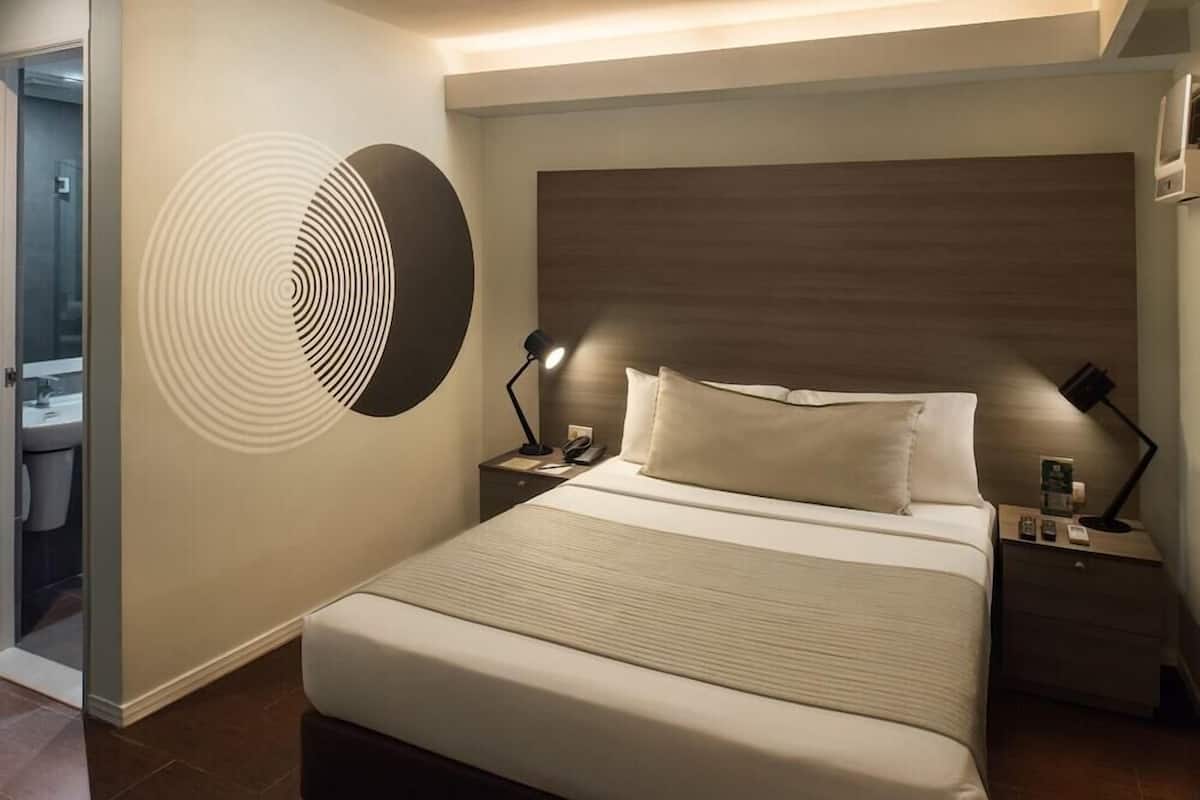 A modern hotel room with a large bed, a wooden headboard, two bedside tables with lamps, and a wall art piece.