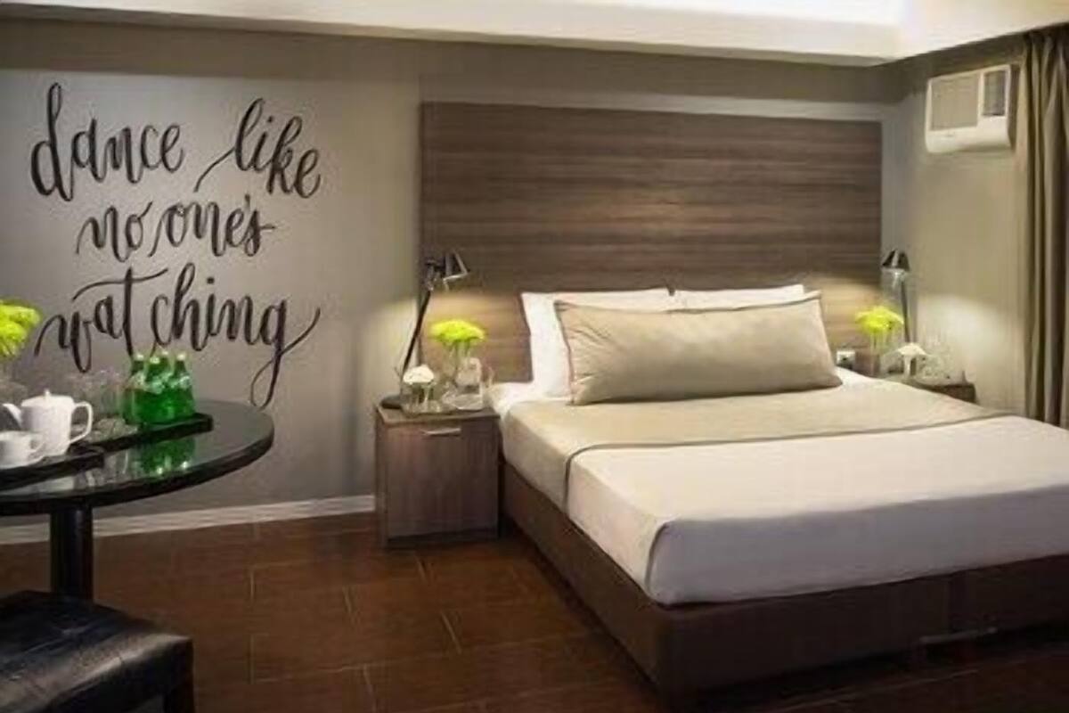 A hotel room with a bed, bedside tables, a small table with a lamp, and a wall with a motivational quote.