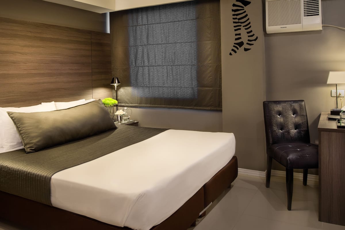 A modern hotel room with a large bed, a desk, a chair, and a window with blinds.