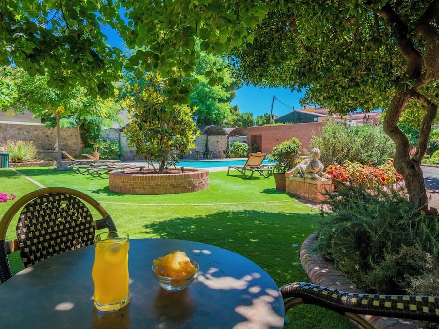 Eine Terrasse mit einem runden Tisch, einem Stuhl und einem Getränk. Im Hintergrund ist ein Pool zu sehen, der von Grünpflanzen und einer Backsteinmauer umgeben ist.