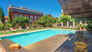 A swimming pool with lounge chairs and a wooden deck, surrounded by greenery and a building in the background.