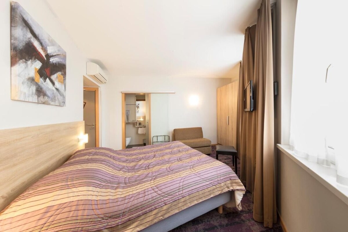 Et hotelværelse med en seng, en stol, et skrivebord og et vindue med gardiner.