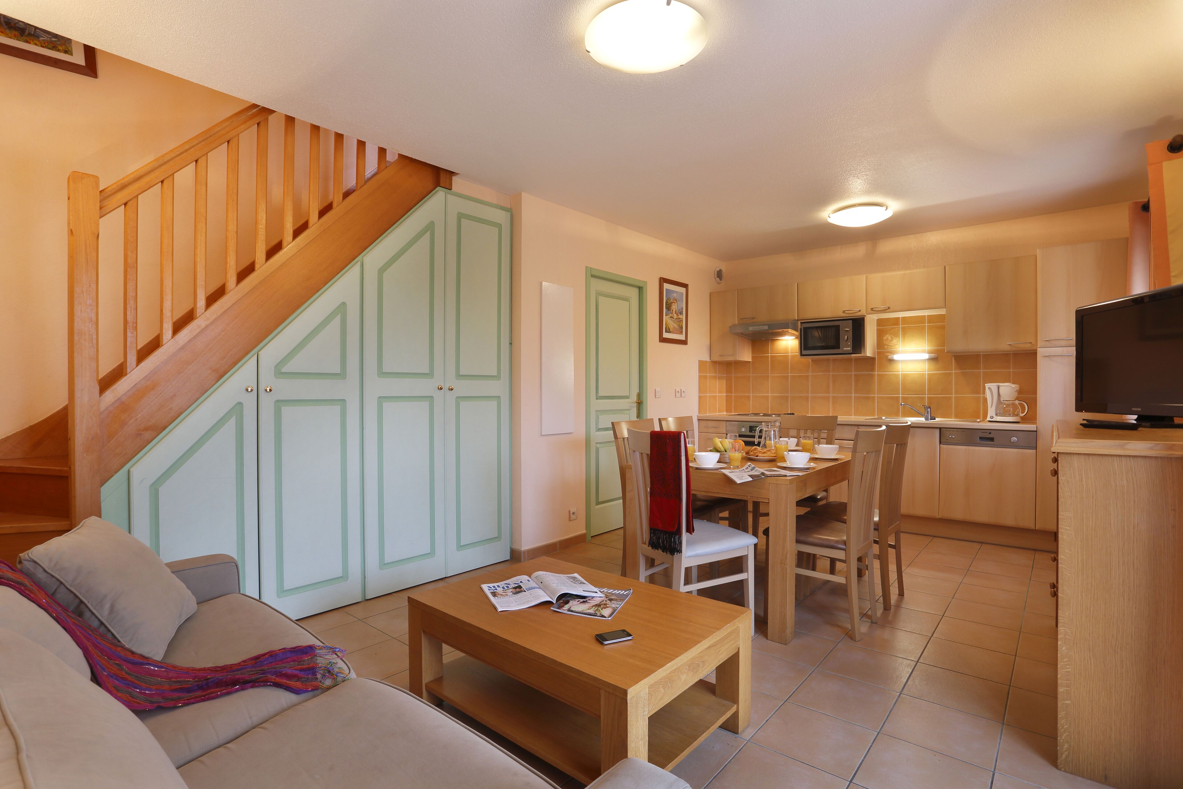 A modern kitchen with wooden cabinets, a dining area with a table and chairs, and a staircase leading to another level.