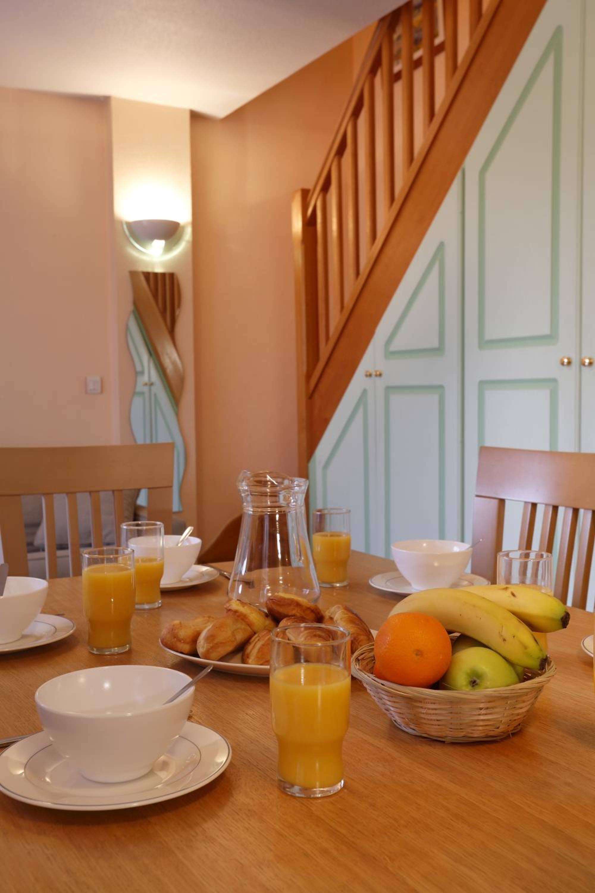 Una mesa de desayuno con platos, vasos, una jarra y una cesta de frutas y pasteles.