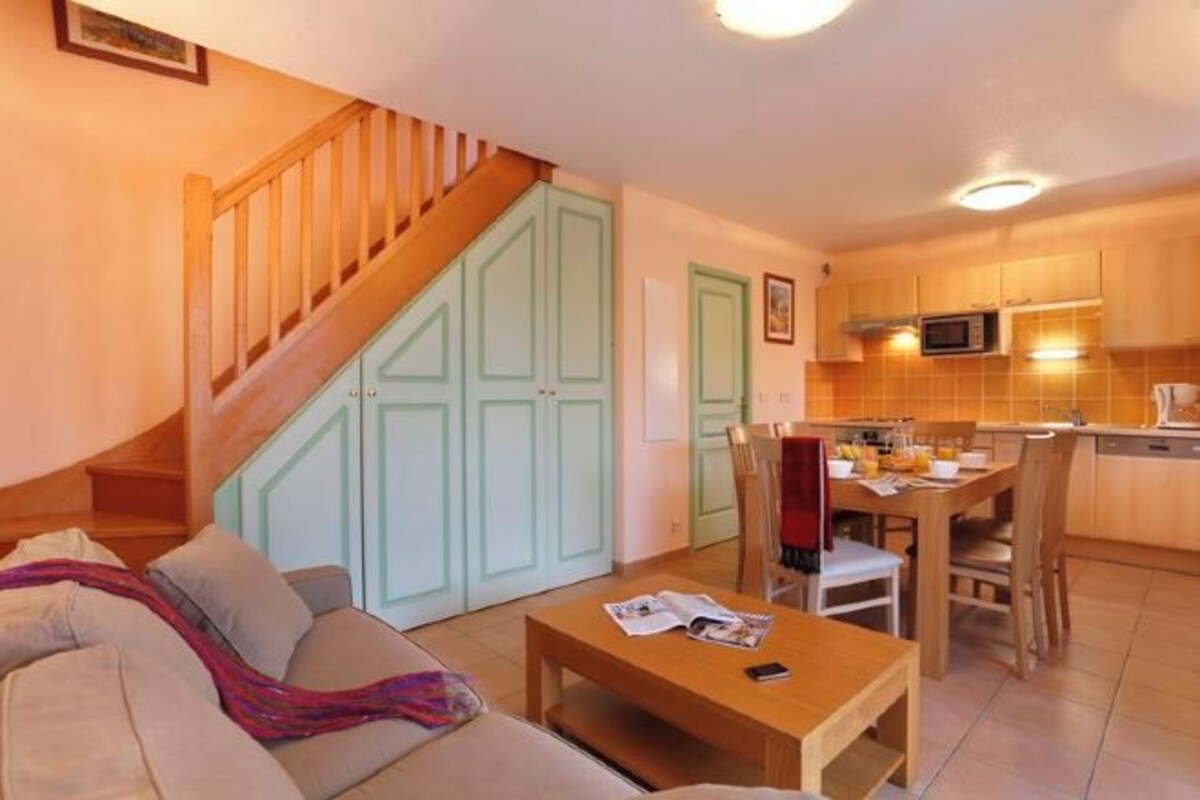 Un salon comprenant un canapé, une table basse et un coin repas avec des chaises et une table. Un escalier mène à une porte verte.
