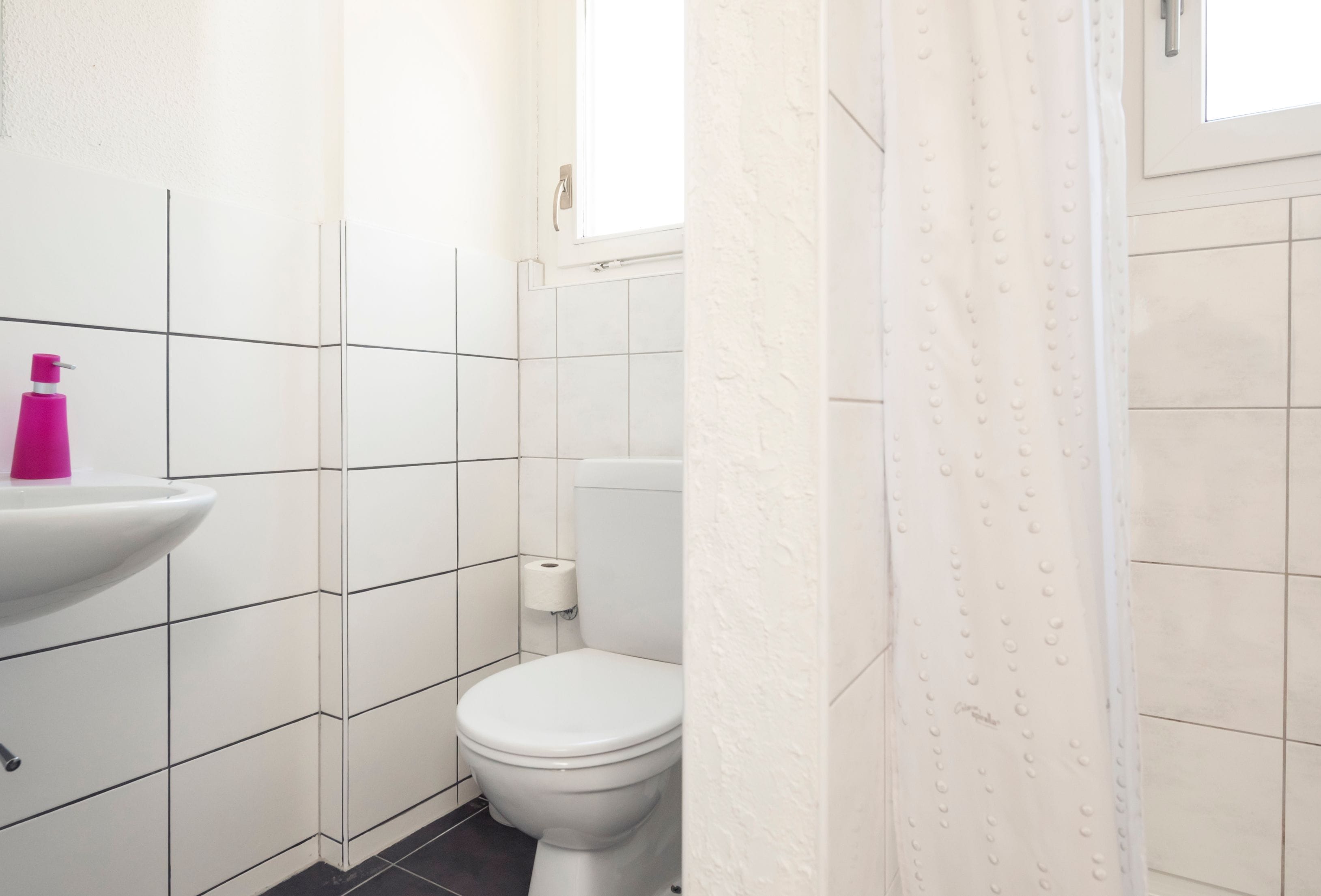 A bathroom with white tiled walls, a white sink with a pink soap dispenser, a toilet, and a shower curtain.