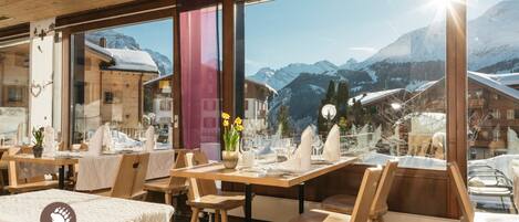 A restaurant with wooden tables and chairs, large windows offering mountain views, and a sign reading "Bären Restaurant".