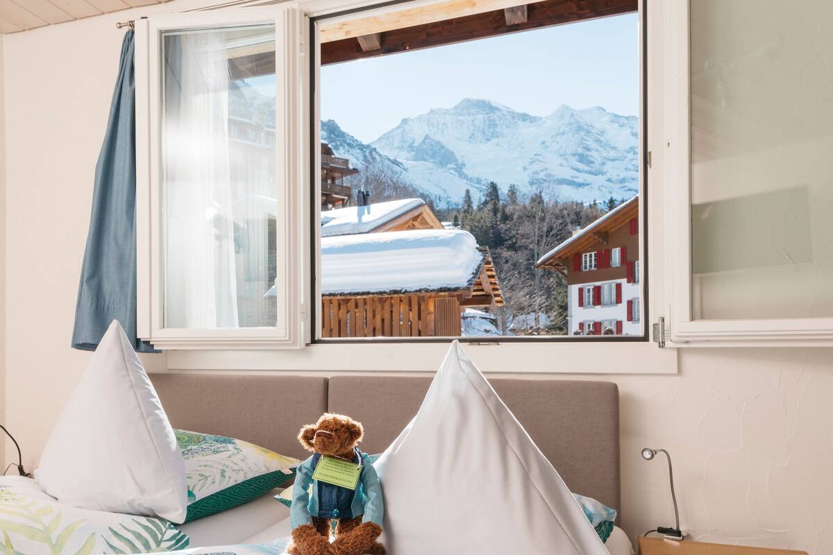Un lit avec un ours en peluche, une fenêtre donnant sur les montagnes et une cabane en bois.