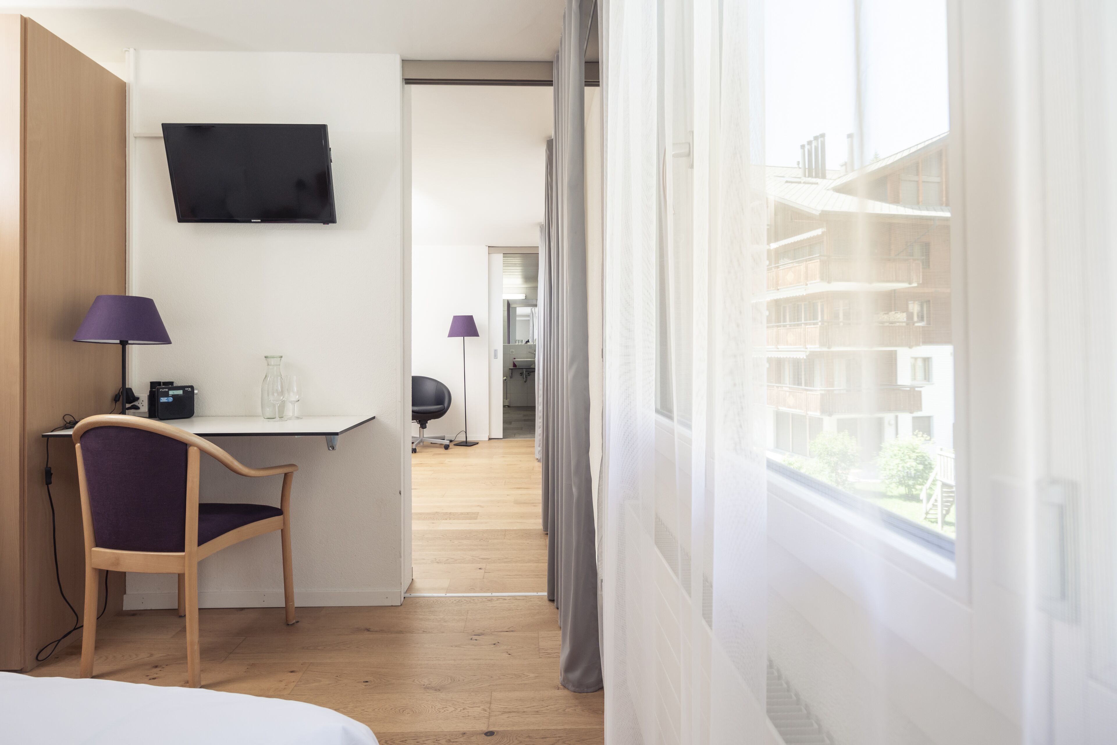 Ein Hotelzimmer mit Bett, Schreibtisch mit Stuhl, einem an der Wand montierten Fernseher und Blick auf Balkon.