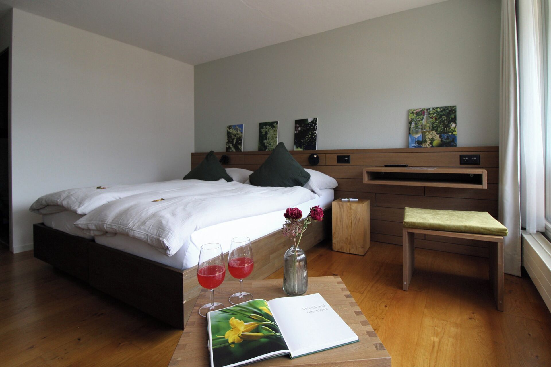 Habitación de hotel moderna con cama, mesita de noche y escritorio con silla. Hay dos cuadros en la pared, un jarrón con flores y un libro sobre la mesa.