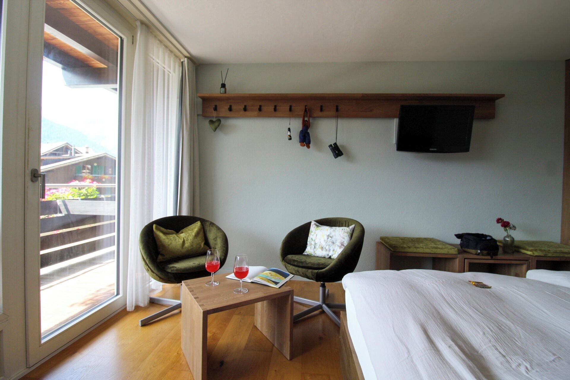 A hotel room with a bed, two chairs, a small table with drinks, a TV mounted on the wall, and a window with curtains.