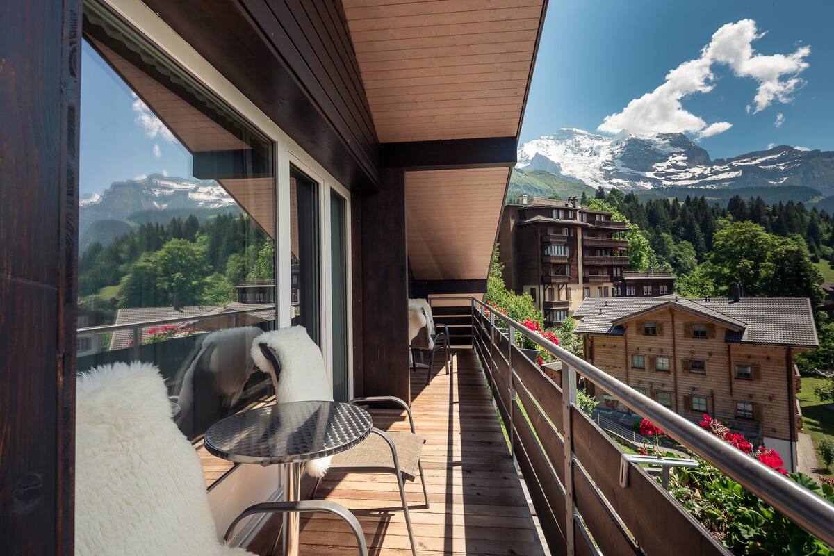 Un balcon offrant une vue sur des montagnes enneigées, des bâtiments en bois et une chaise.
