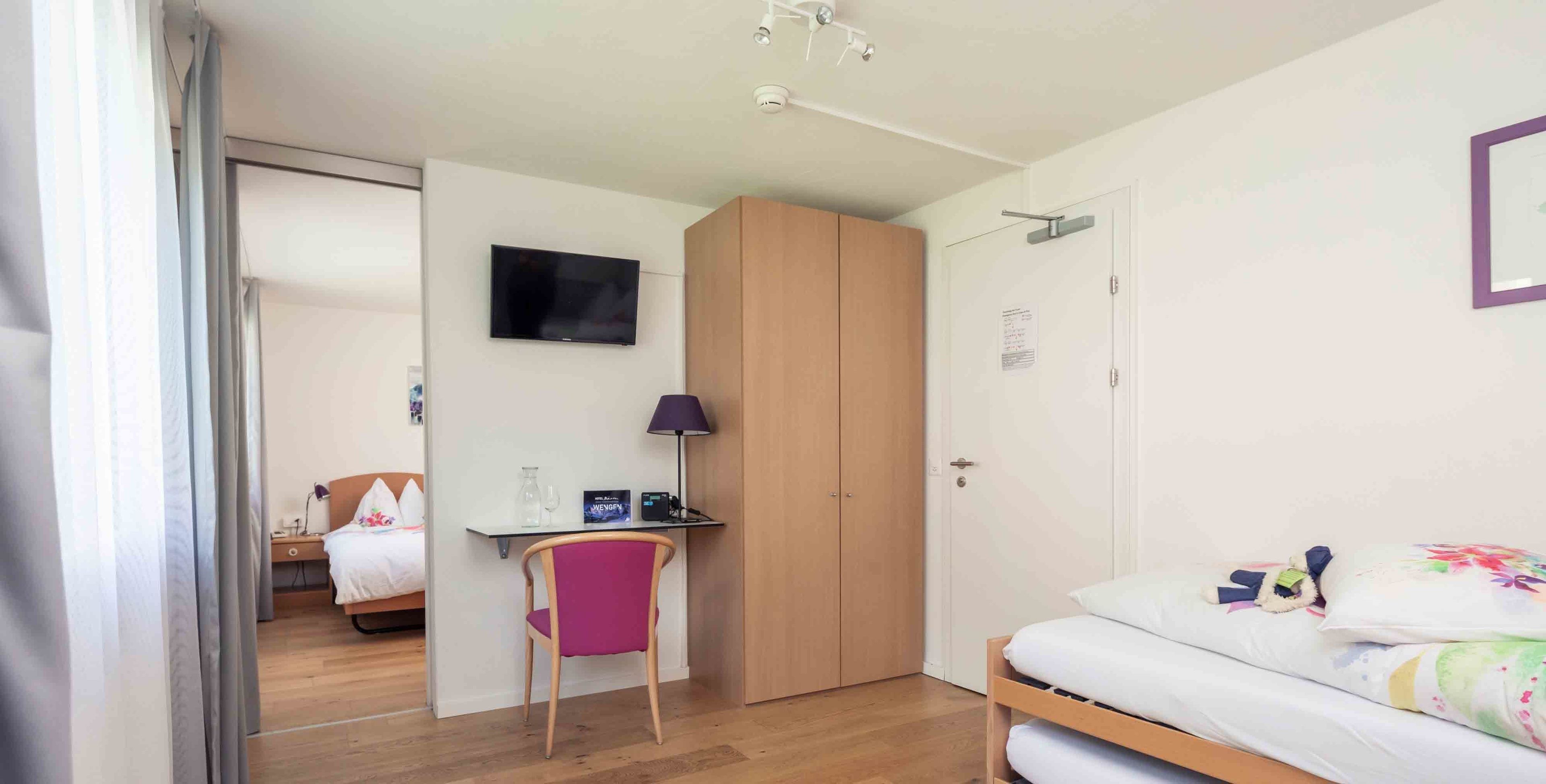 A bedroom with a bed, a desk with a chair, a wardrobe, and a television on the wall.
