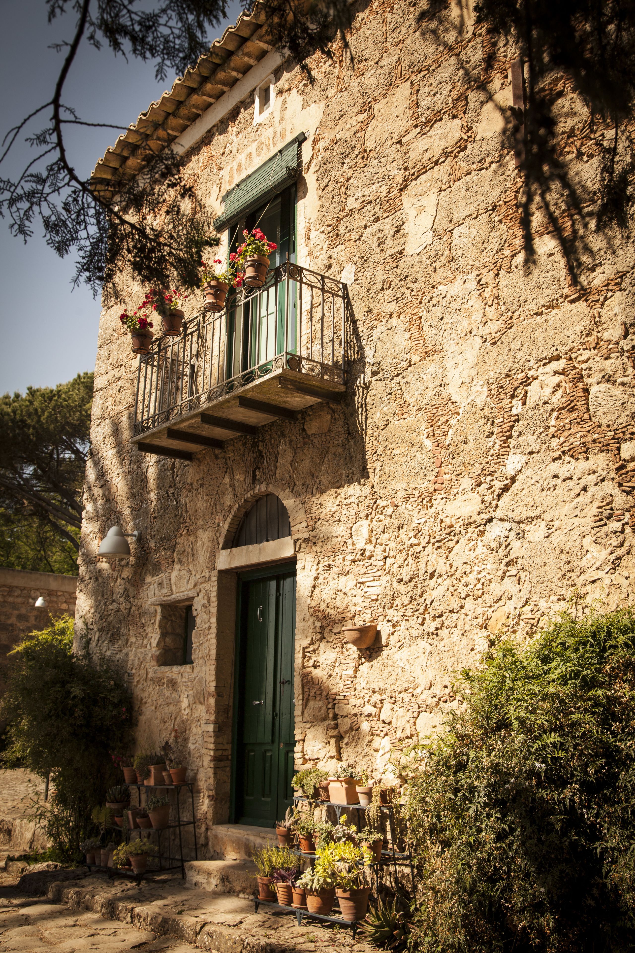A rustic stone building with a green door, a balcony with potted plants, and a small window.
