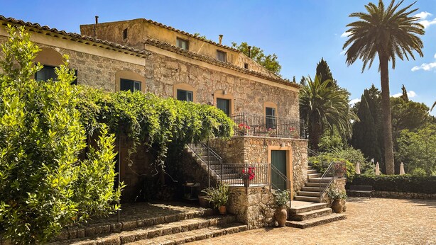 A stone building with a balcony, steps leading to a door, and a palm tree in the background. - Masseria Bannata (Enna)