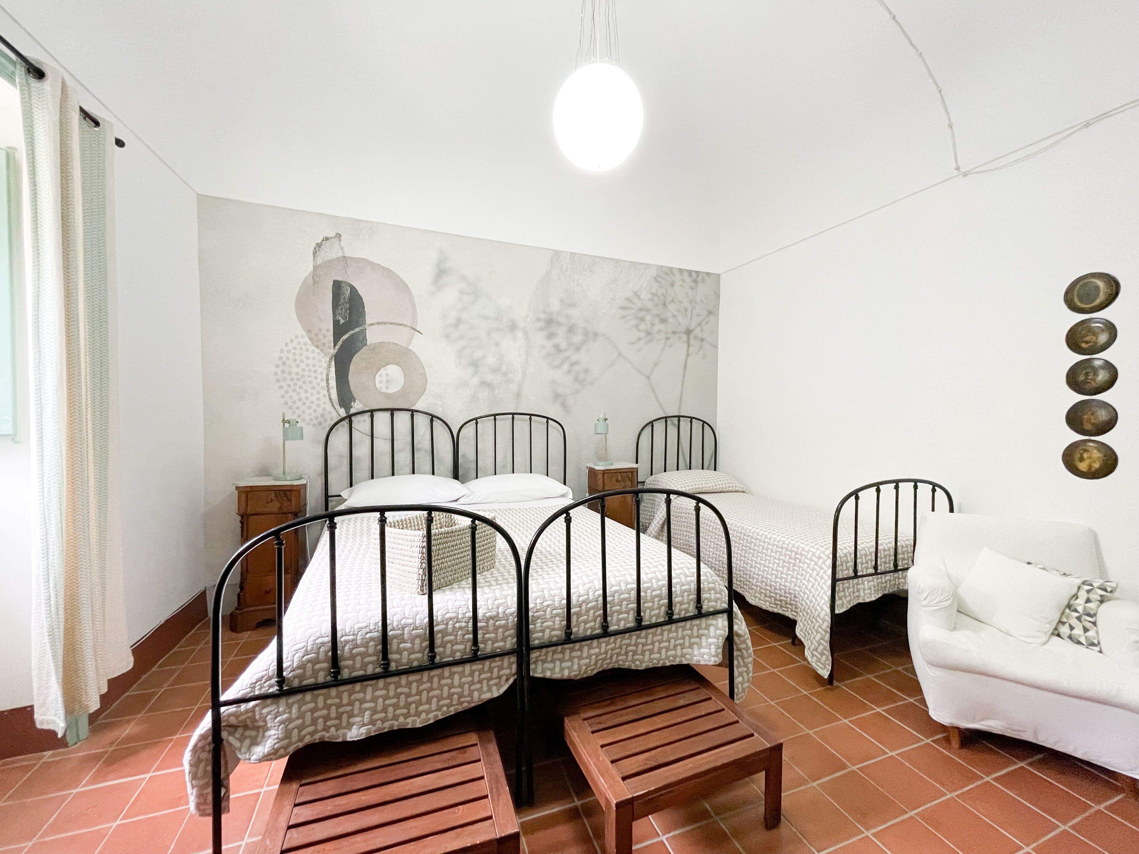 Una habitación con dos camas, un sofá blanco, un banco de madera, una pared con arte abstracto e iluminación en el techo.