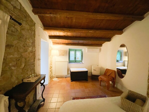 A cozy bedroom with a stone wall, a wooden ceiling, a bed, a chair, a small table, and a window with green shutters.