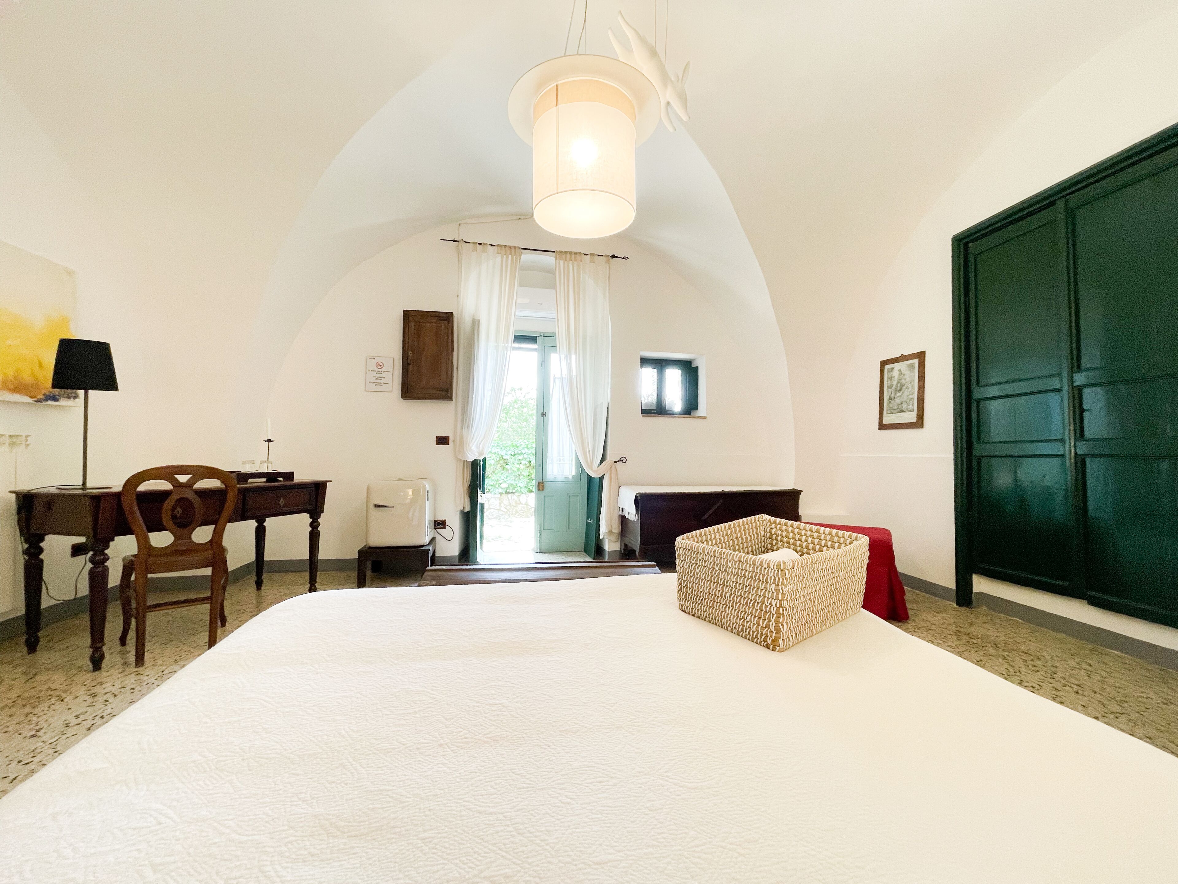 A room with a large bed, a desk with a chair, a lamp, and a basket.