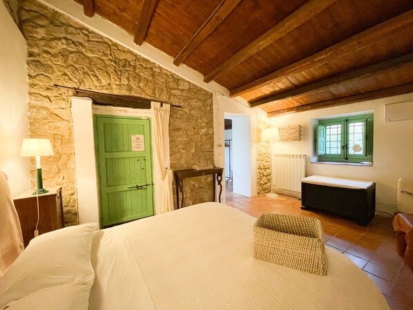 A bedroom with a stone wall, a green door, a bed, a bench, and a window with shutters.