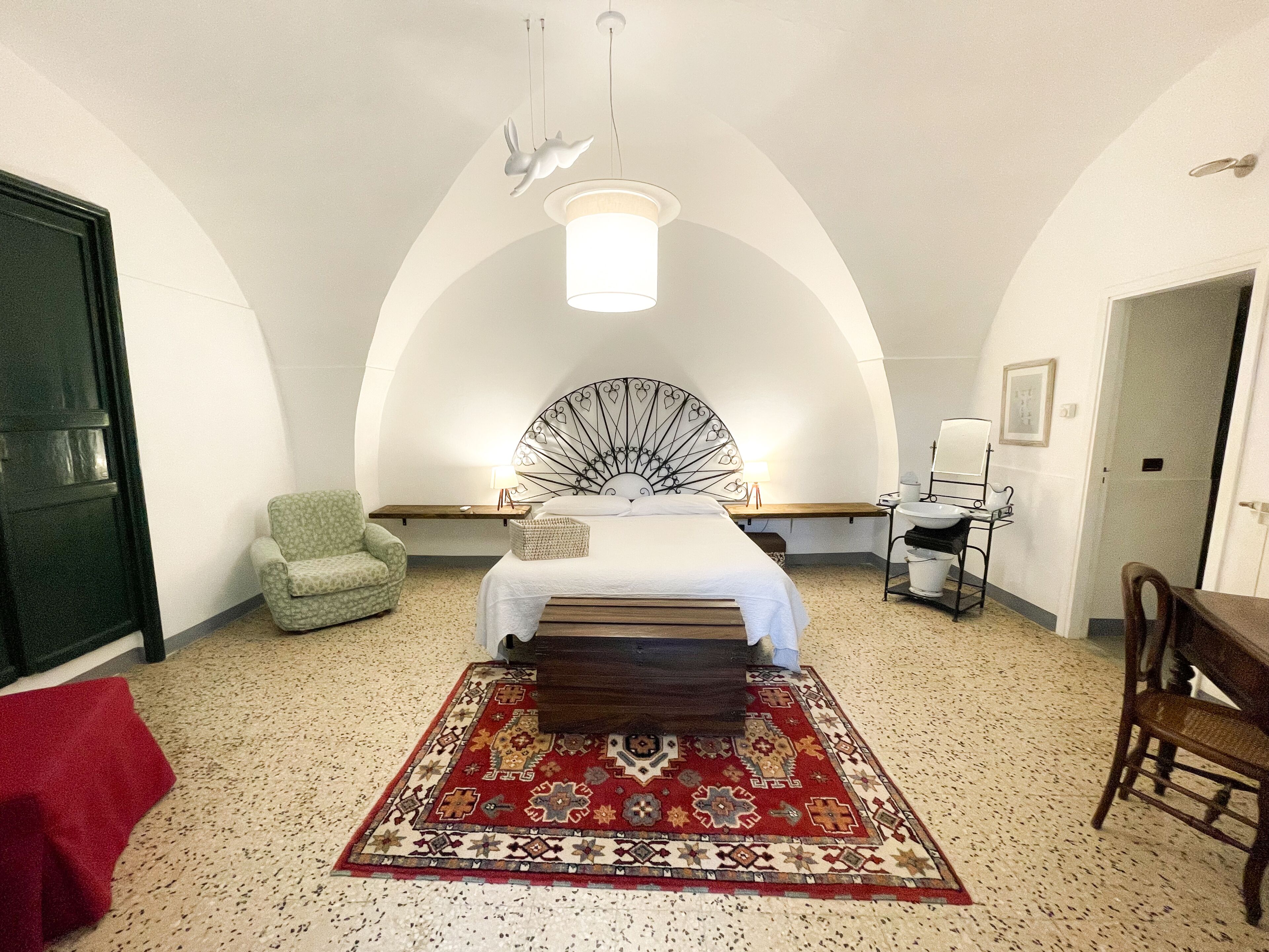 Ein Schlafzimmer mit einem Bett, einem grünen Sessel, einer Holzbank, einem roten Teppich mit Muster, einer weißen Wand, einer weißen Decke und einer Pendelleuchte.