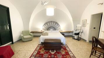 Ein Schlafzimmer mit einem Bett, einem grünen Sessel, einer Holzbank, einem roten Teppich mit Muster, einer weißen Wand, einer weißen Decke und einer Pendelleuchte.