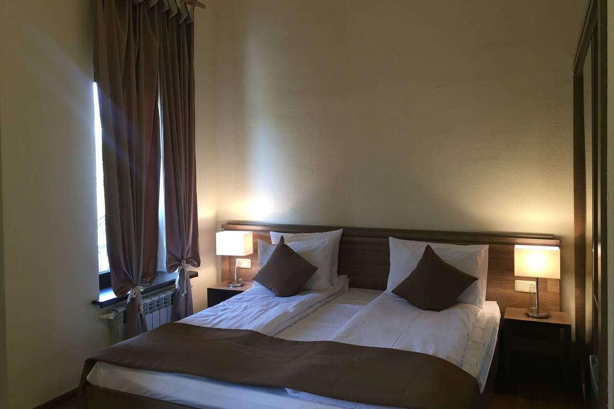 A hotel room with a large bed, two bedside tables with lamps, a window with curtains, and a radiator.