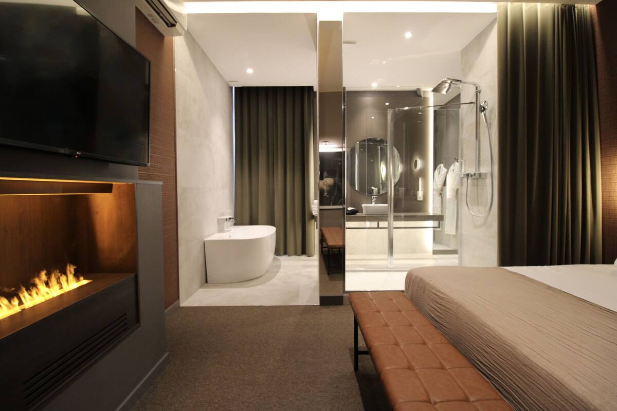 A modern hotel room with a flat-screen TV, a fireplace, a large bed, a bench, a bathroom with a shower, and a glass-enclosed shower area.